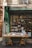 A quaint bookstore facade with a green awning, featuring a wooden table outside displaying a variety of books. The glass window behind showcases more books and has a warm interior light illuminating the shop.
