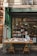 A quaint bookstore facade with a green awning, featuring a wooden table outside displaying a variety of books. The glass window behind showcases more books and has a warm interior light illuminating the shop.