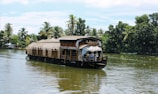A traditional houseboat with a thatched roof is gently floating on a calm river surrounded by lush green palm trees. The boat features intricate wooden architecture and has a small covered deck at the front.