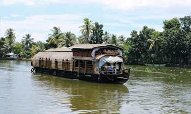 A traditional houseboat with a thatched roof is gently floating on a calm river surrounded by lush green palm trees. The boat features intricate wooden architecture and has a small covered deck at the front.