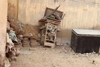 A dusty courtyard corner with a stack of wooden pallets and old furniture, including a broken chair and table. Burlap sacks and rough bricks are piled on one side, and a large black metal box is on the other, surrounded by small debris and stones. The walls are weathered and slightly peeling.