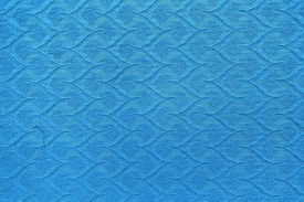 A close-up view of a textured blue surface with a repeating wave-like pattern. The intricate design features a series of diamond-shaped elements that interlock, creating a sense of movement across the fabric.