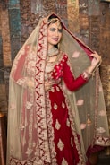 A smiling bride wearing a traditional red and gold embroidered bridal outfit, including an elaborate necklace and a detailed headpiece, stands in front of a decorative wooden backdrop. The bride is lifting the veil slightly with one hand, showing her face and jewelry.