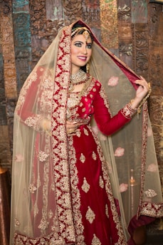 A smiling bride wearing a traditional red and gold embroidered bridal outfit, including an elaborate necklace and a detailed headpiece, stands in front of a decorative wooden backdrop. The bride is lifting the veil slightly with one hand, showing her face and jewelry.