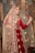 A smiling bride wearing a traditional red and gold embroidered bridal outfit, including an elaborate necklace and a detailed headpiece, stands in front of a decorative wooden backdrop. The bride is lifting the veil slightly with one hand, showing her face and jewelry.