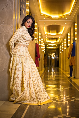 A woman in an elegant, patterned gown stands against a marble wall in a luxurious, well-lit hallway. The hallway is adorned with ornate lighting fixtures and reflective floors, creating a warm, golden ambiance.