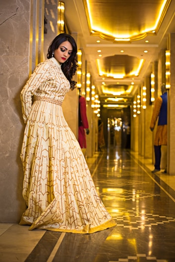 A woman in an elegant, patterned gown stands against a marble wall in a luxurious, well-lit hallway. The hallway is adorned with ornate lighting fixtures and reflective floors, creating a warm, golden ambiance.
