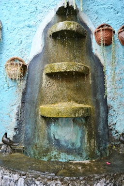 A tiered stone fountain with water trickling down its moss-covered surfaces is mounted on a textured blue wall. Clay pots with trailing plants are hung on either side, adding a rustic and natural touch.