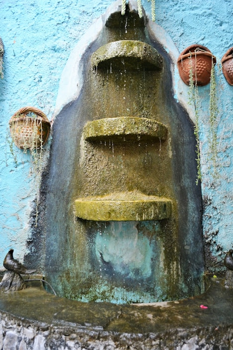 A tiered stone fountain with water trickling down its moss-covered surfaces is mounted on a textured blue wall. Clay pots with trailing plants are hung on either side, adding a rustic and natural touch.