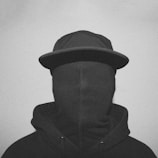 A person wearing a hooded sweatshirt and a cap with their face completely covered by a fabric material, creating a mysterious and anonymous appearance.