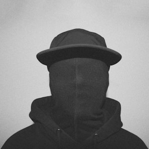 A person wearing a hooded sweatshirt and a cap with their face completely covered by a fabric material, creating a mysterious and anonymous appearance.