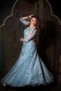 A woman with long brown hair and a bright smile is wearing an elegant light blue gown decorated with floral patterns. She is standing against a dark, intricately designed wooden background, posing with one hand in her hair and the other on her hip.