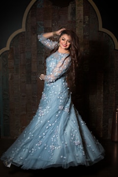 A woman with long brown hair and a bright smile is wearing an elegant light blue gown decorated with floral patterns. She is standing against a dark, intricately designed wooden background, posing with one hand in her hair and the other on her hip.