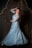A woman with long brown hair and a bright smile is wearing an elegant light blue gown decorated with floral patterns. She is standing against a dark, intricately designed wooden background, posing with one hand in her hair and the other on her hip.