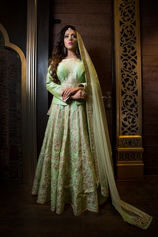 A woman stands elegantly in a richly embroidered mint green traditional dress with intricate gold patterns. Her long, dark hair cascades over her shoulders, and she wears a matching dupatta draped over her head. The setting features ornate, intricately carved wooden panels, adding to the luxurious feel.