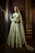A woman stands elegantly in a richly embroidered mint green traditional dress with intricate gold patterns. Her long, dark hair cascades over her shoulders, and she wears a matching dupatta draped over her head. The setting features ornate, intricately carved wooden panels, adding to the luxurious feel.