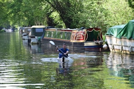 A person is kayaking on a calm canal lined with moored boats, surrounded by lush green trees. The sunlight filters through the foliage, casting a mix of light and shadows on the water's surface. Some of the boats have vibrant colors and appear to be houseboats or barges, adding a quaint charm to the scene.