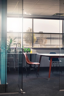 An office space with a large window allowing natural light to illuminate the room. Inside, there are modern chairs around a rectangular table. Green plants decorate the corner, and papers sit on the table, suggesting minimal use. The floor is carpeted, and the walls are glass, providing a view of the exterior.