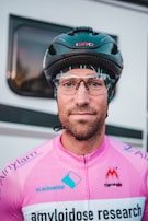 A person wearing a black cycling helmet and clear glasses stands facing the camera. They are dressed in a bright pink cycling jersey with various logos, including 'amyloidose research'. The background appears to be an indistinct building with rectangular windows.
