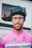 A person wearing a black cycling helmet and clear glasses stands facing the camera. They are dressed in a bright pink cycling jersey with various logos, including 'amyloidose research'. The background appears to be an indistinct building with rectangular windows.