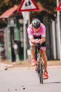 A cyclist wearing a pink jersey and helmet rides a bicycle on a street. The cyclist is in a racing position, with focus and intensity visible. Traffic signs are slightly out of focus in the background, indicating a road environment.
