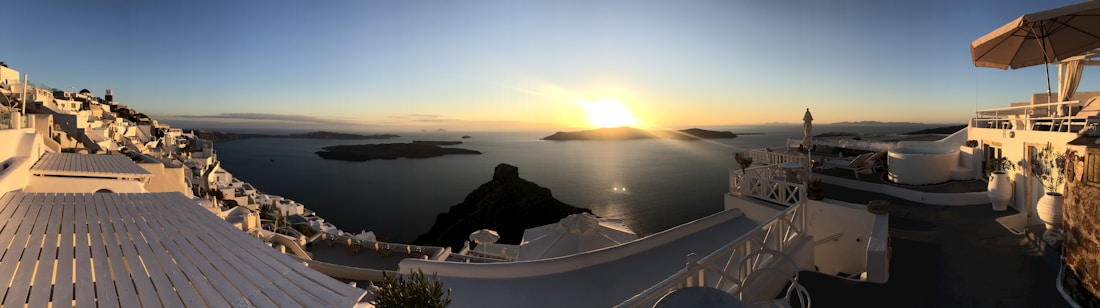 A picturesque coastal view at sunset features whitewashed buildings with blue accents cascading down a hillside towards the sea. The sun is setting over the horizon, casting a warm golden glow across the water and the buildings. Several small islands are visible in the distance, surrounded by the deep blue of the ocean.