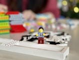 A LEGO minifigure is standing on a white LEGO base with blurred colorful LEGO blocks in the background. The figure is wearing a blue shirt, red tie, and red pants. The background is out of focus, creating a soft visual effect.