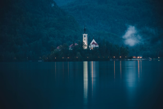 A serene lakeside scene during twilight with a small island featuring a prominent church and steeple surrounded by dense forests. The calm water reflects the lights from the island, while mist gently hovers over the trees.