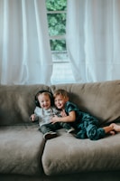 Two young children are sitting together on a beige couch in a cozy environment. One child is wearing headphones and a light-colored shirt while the other is dressed in blue pajamas. They are smiling and seem to be enjoying each other's company. Behind them, large windows with sheer white curtains allow light to illuminate the room.