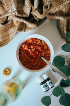 A white bowl filled with a red, textured substance possibly resembling a spicy sauce or stew, accompanied by a spoon. A plaid brown fabric is draped in the background. Two small bottles with labels are placed nearby, along with a bottle cap on the left, suggesting the presence of a beverage. Green leaves are arranged on the right side, adding a natural element to the composition.