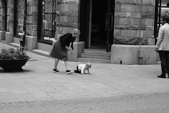 A black and white street scene features a woman bending over to assist a child who is crawling on the pavement. The setting is urban, with storefronts visible in the background. There is a large planter to the left and a man standing to the right, observing the interaction.