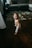 A young child wearing diapers is standing on a wooden floor, holding a remote control. The room features a gray couch with pillows and a patterned rug. Toys and other items are scattered on the floor, giving a cozy and lived-in atmosphere.