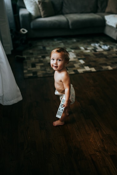 A young child wearing diapers is standing on a wooden floor, holding a remote control. The room features a gray couch with pillows and a patterned rug. Toys and other items are scattered on the floor, giving a cozy and lived-in atmosphere.