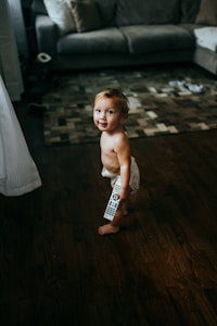 A young child wearing diapers is standing on a wooden floor, holding a remote control. The room features a gray couch with pillows and a patterned rug. Toys and other items are scattered on the floor, giving a cozy and lived-in atmosphere.