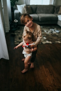 A young child is gently holding a toddler who is wearing a diaper and holding a small red toy. They are standing on a wood floor in a cozy living room with a gray couch and a patterned rug in the background. Sunlight filters through a nearby window, casting soft shadows.