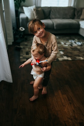 A young child is gently holding a toddler who is wearing a diaper and holding a small red toy. They are standing on a wood floor in a cozy living room with a gray couch and a patterned rug in the background. Sunlight filters through a nearby window, casting soft shadows.