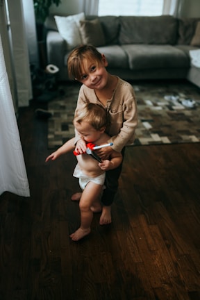 A young child is gently holding a toddler who is wearing a diaper and holding a small red toy. They are standing on a wood floor in a cozy living room with a gray couch and a patterned rug in the background. Sunlight filters through a nearby window, casting soft shadows.