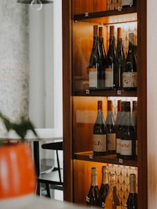 A wooden wine rack with shelves displaying multiple bottles of wine. The background appears to have a decorative pattern, and the scene is warmly lit. In the foreground, a blurred element suggests a modern and cozy interior setting, possibly part of a restaurant or bar.