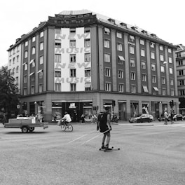 A busy urban intersection with a multi-story building featuring large text advertising 'New Music.' People are engaged in various activities including a person on a bicycle, another on roller skis, and pedestrians walking along the sidewalk. The building has a storefront for Adidas, and there are vehicles on the road including a car and a small trailer.