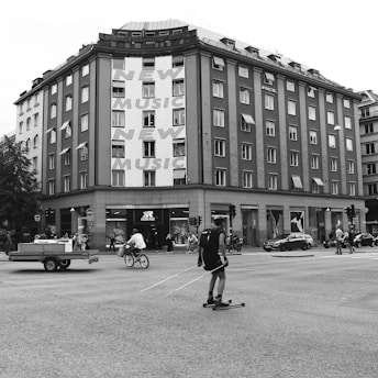 A busy urban intersection with a multi-story building featuring large text advertising 'New Music.' People are engaged in various activities including a person on a bicycle, another on roller skis, and pedestrians walking along the sidewalk. The building has a storefront for Adidas, and there are vehicles on the road including a car and a small trailer.