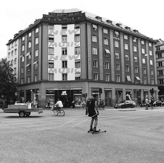 A busy urban intersection with a multi-story building featuring large text advertising 'New Music.' People are engaged in various activities including a person on a bicycle, another on roller skis, and pedestrians walking along the sidewalk. The building has a storefront for Adidas, and there are vehicles on the road including a car and a small trailer.