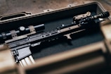A tactical rifle placed inside a sturdy, foam-lined case. The firearm features various attachments, including a scope and a flashlight, and exhibits a blend of black and tan colors.