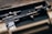 A tactical rifle placed inside a sturdy, foam-lined case. The firearm features various attachments, including a scope and a flashlight, and exhibits a blend of black and tan colors.