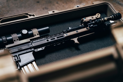 A tactical rifle placed inside a sturdy, foam-lined case. The firearm features various attachments, including a scope and a flashlight, and exhibits a blend of black and tan colors.