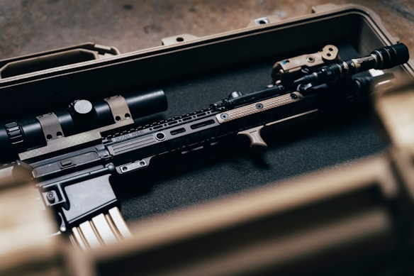 A tactical rifle placed inside a sturdy, foam-lined case. The firearm features various attachments, including a scope and a flashlight, and exhibits a blend of black and tan colors.