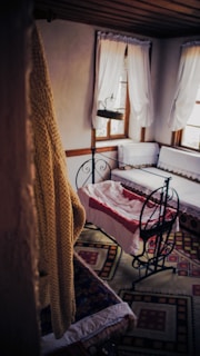 A cozy, vintage-style room features a cradle with a red and white blanket. The room is decorated with ornate rugs on the floor, white curtains drape over two windows, and a knitted mustard-colored blanket hangs in the foreground.