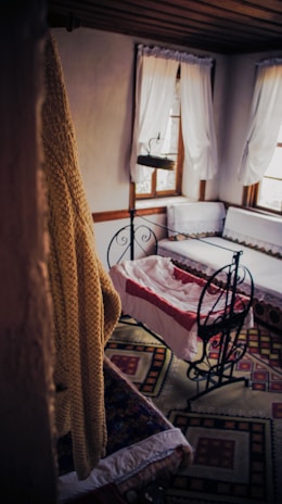 A cozy, vintage-style room features a cradle with a red and white blanket. The room is decorated with ornate rugs on the floor, white curtains drape over two windows, and a knitted mustard-colored blanket hangs in the foreground.