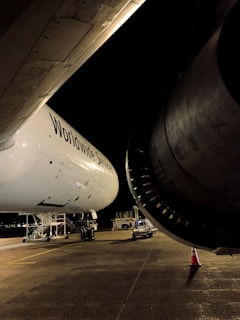 A large airplane is parked on an airport tarmac under dim lighting. The aircraft's engine and wing are prominently visible. Ground service vehicles and equipment are stationed nearby, and a traffic cone is placed in front of the engine for safety.
