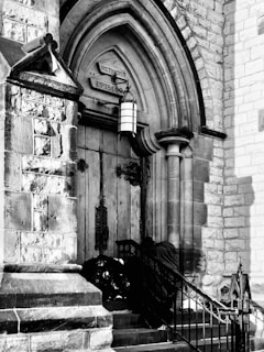 The entrance of a historic church featuring ornate wooden doors and stone archways. Above the door, a sign reads 'Methodist Episcopal'. Wrought iron railings border the steps leading to the entrance. A person wrapped in a dark fabric is sitting or leaning against the railing near the doors.