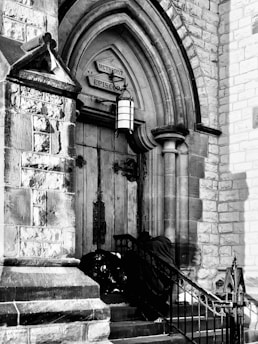 The entrance of a historic church featuring ornate wooden doors and stone archways. Above the door, a sign reads 'Methodist Episcopal'. Wrought iron railings border the steps leading to the entrance. A person wrapped in a dark fabric is sitting or leaning against the railing near the doors.
